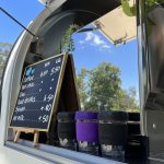 Menu blackboard and three glass keep-cups in a row in front of coffee machine reflecting blue sky and trees