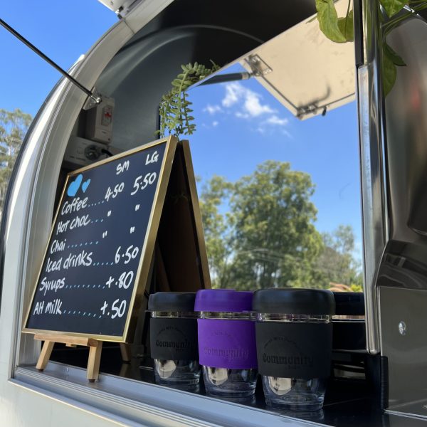 Menu blackboard and three glass keep-cups in a row in front of coffee machine reflecting blue sky and trees